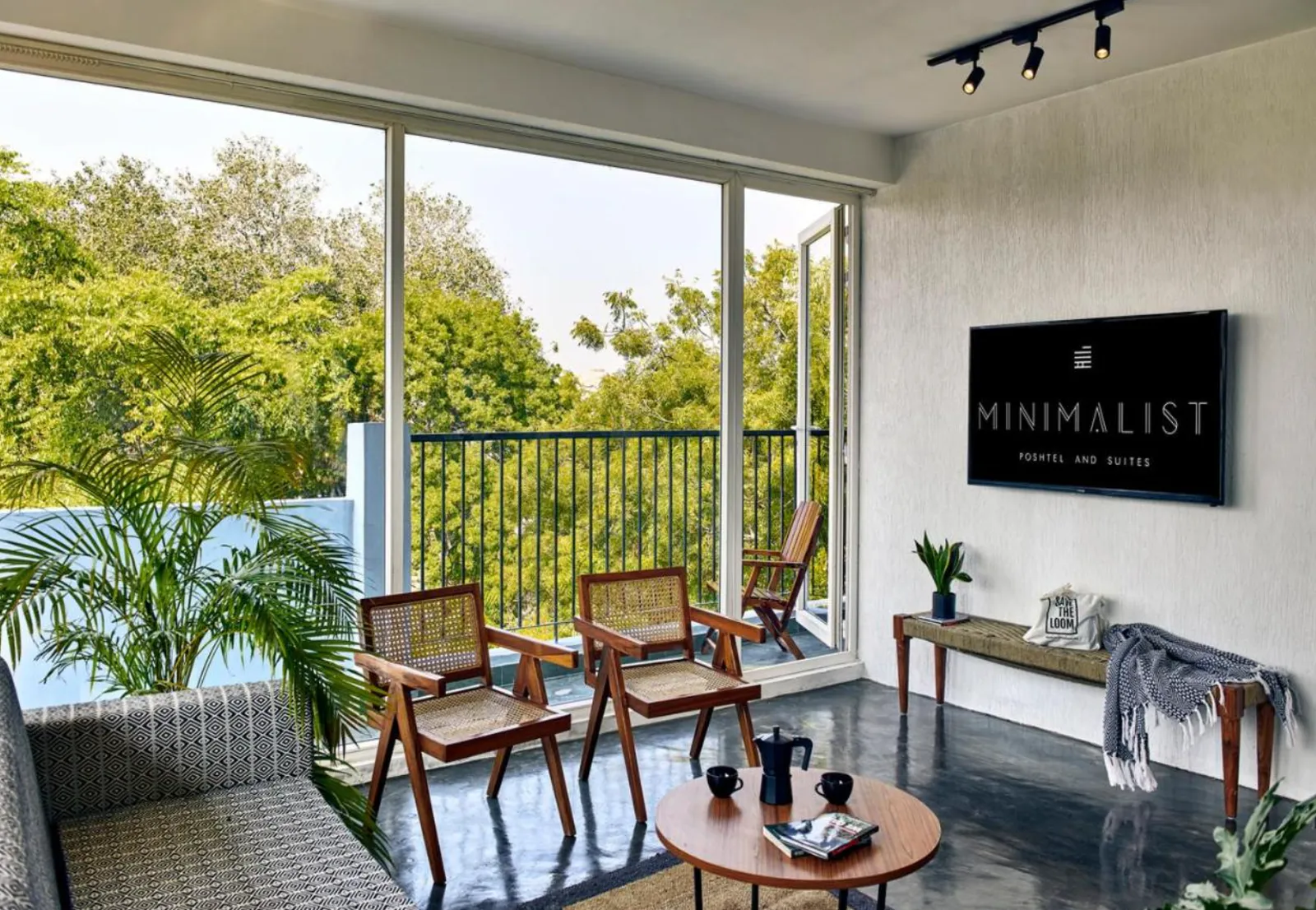 Living room with cane chairs, balcony view of trees, and a TV showing the Minimalist Poshtel and Suites logo