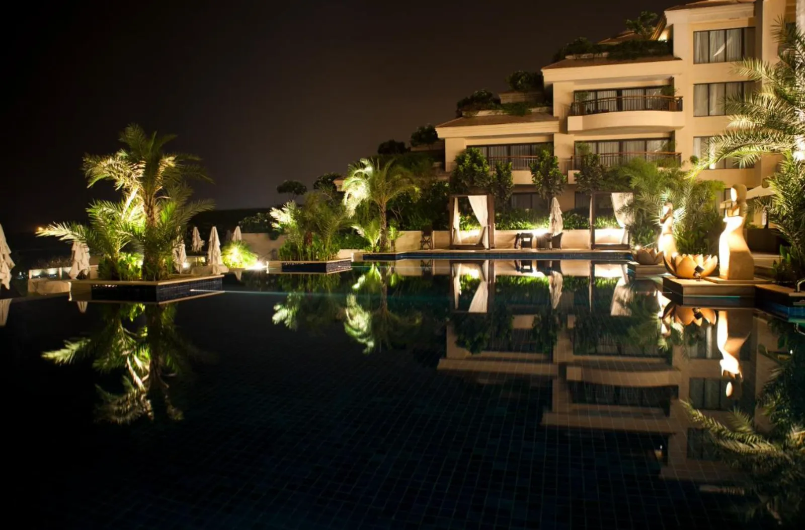 Infinity pool lit at night with palm trees, cabanas and statues reflecting in water